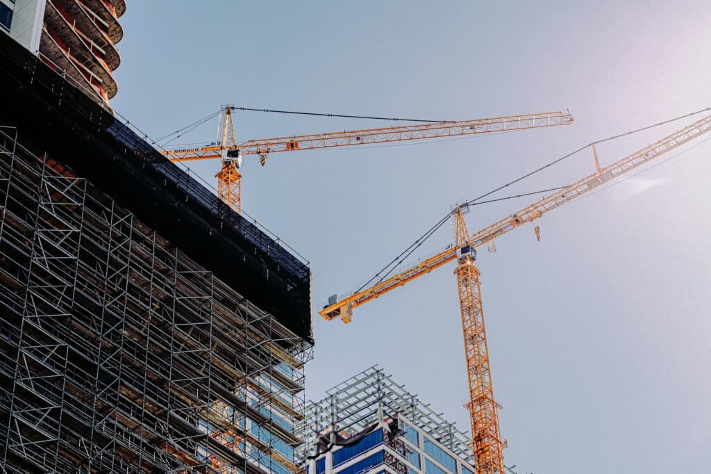 Two large yellow construction cranes operate above partially constructed buildings against a clear blue sky.