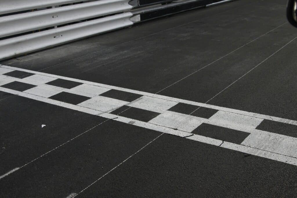 A close-up of a racetrack finish line with a black and white checkered pattern painted across the asphalt.