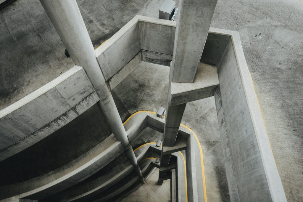 A concrete parking garage structure seen from above, featuring ramps, columns, and yellow lane markings.