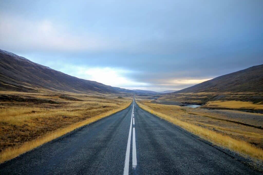 A straight, empty road stretches into the distance through a wide valley with dry grass and hills under a cloudy sky.